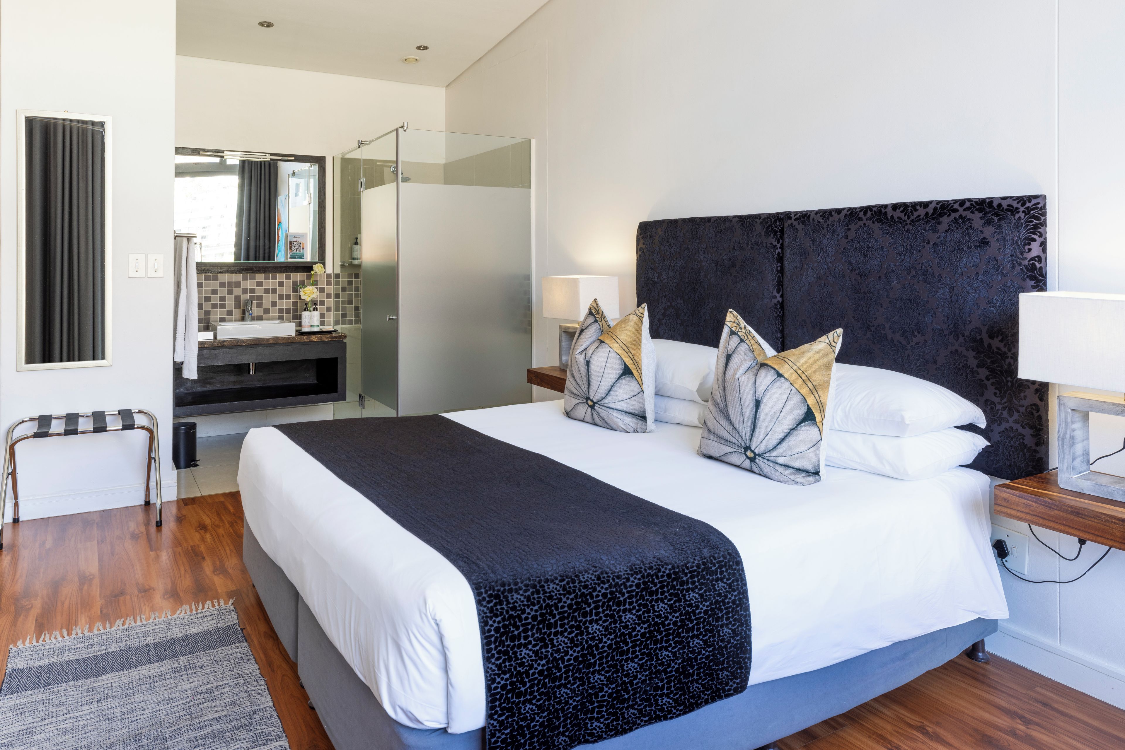 A modern bedroom with a large bed, a bedside table, a nightstand, a mirror, a bathroom with a shower, and a view of the outside.