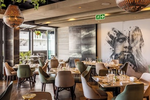 A restaurant interior with tables set for dining, a large abstract mural on the wall, and hanging plants.
