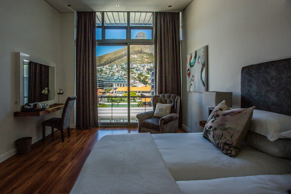 Habitació d'hotel amb un llit gran, una cadira, un escriptori i vista a un paisatge urbà a través d'una finestra gran.