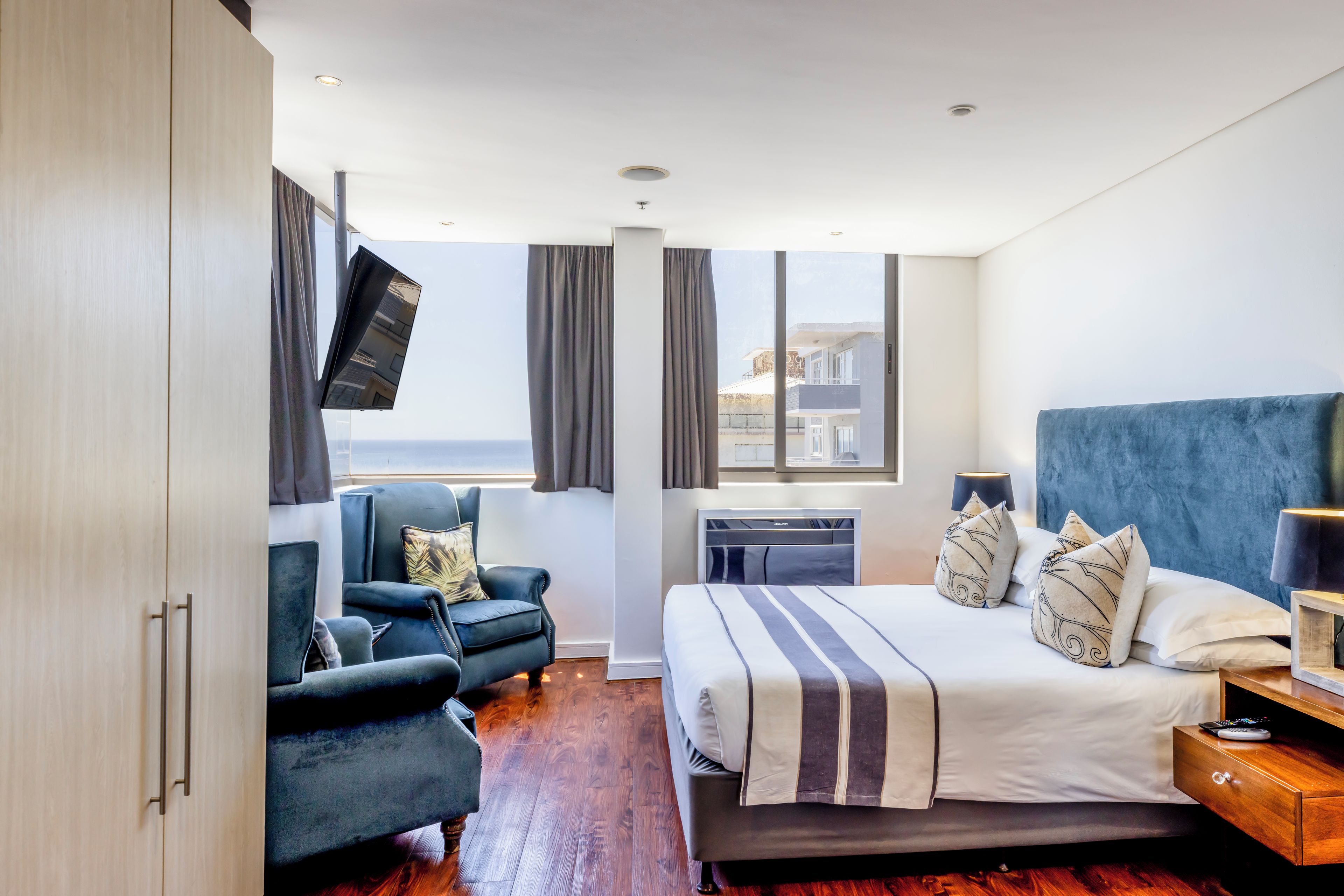 A modern hotel room with a large bed, a seating area with a chair, a desk with a lamp, and a view of the outside.
