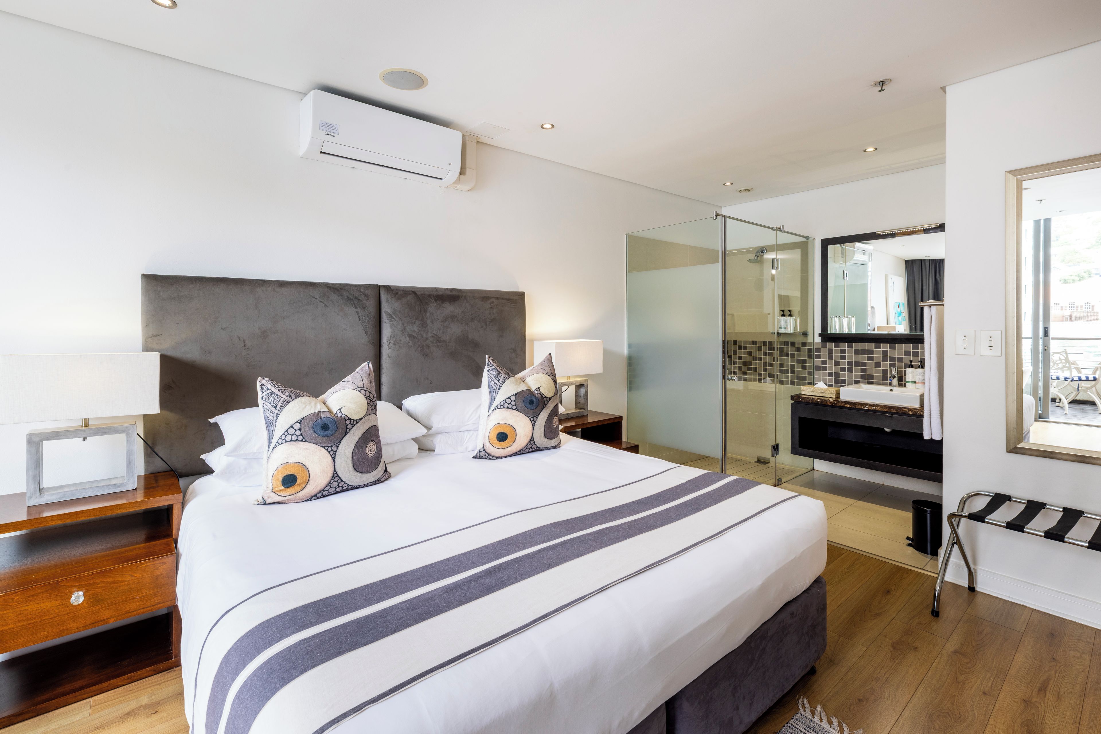 A modern hotel room with a large bed, a glass shower enclosure, a vanity with a mirror, and a view of the outdoors.