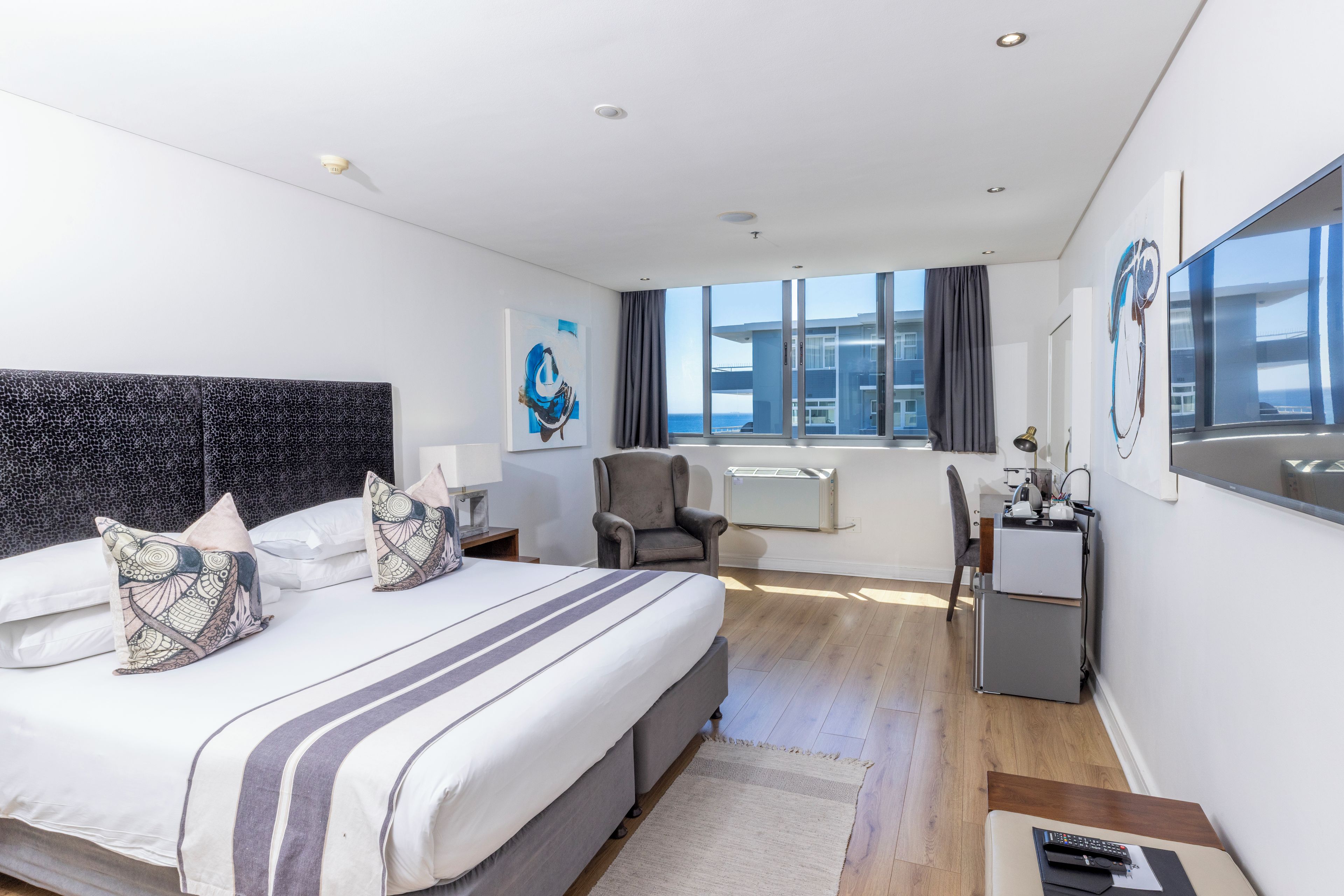 A modern hotel room with a large bed, a desk, a chair, and a view of the city through large windows.