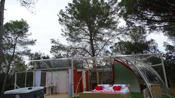 Een transparante tent met een bed en een hottub, omringd door bomen.