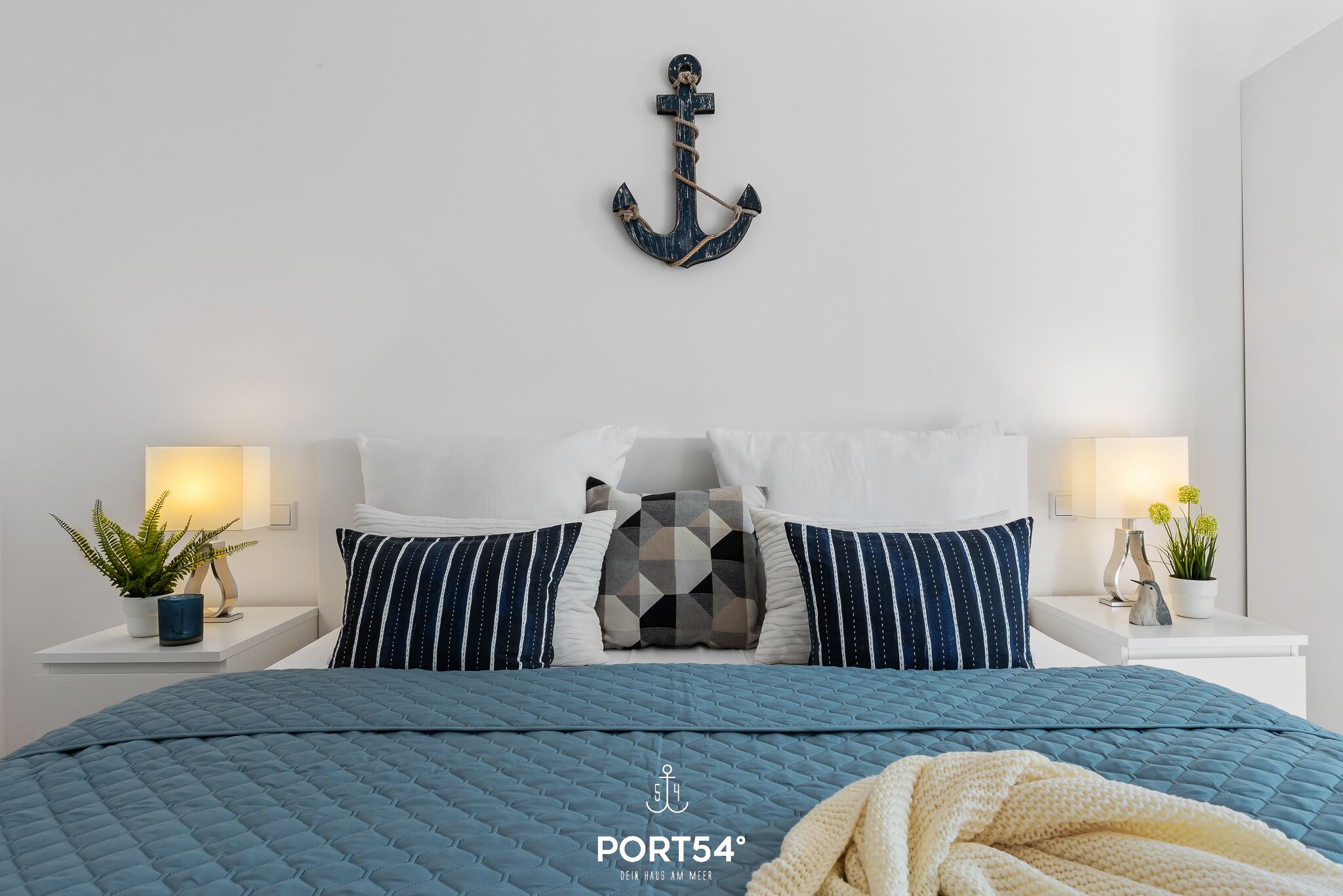 A bedroom with a bed, pillows, bedside tables, a wall-mounted anchor decoration, and a potted plant.