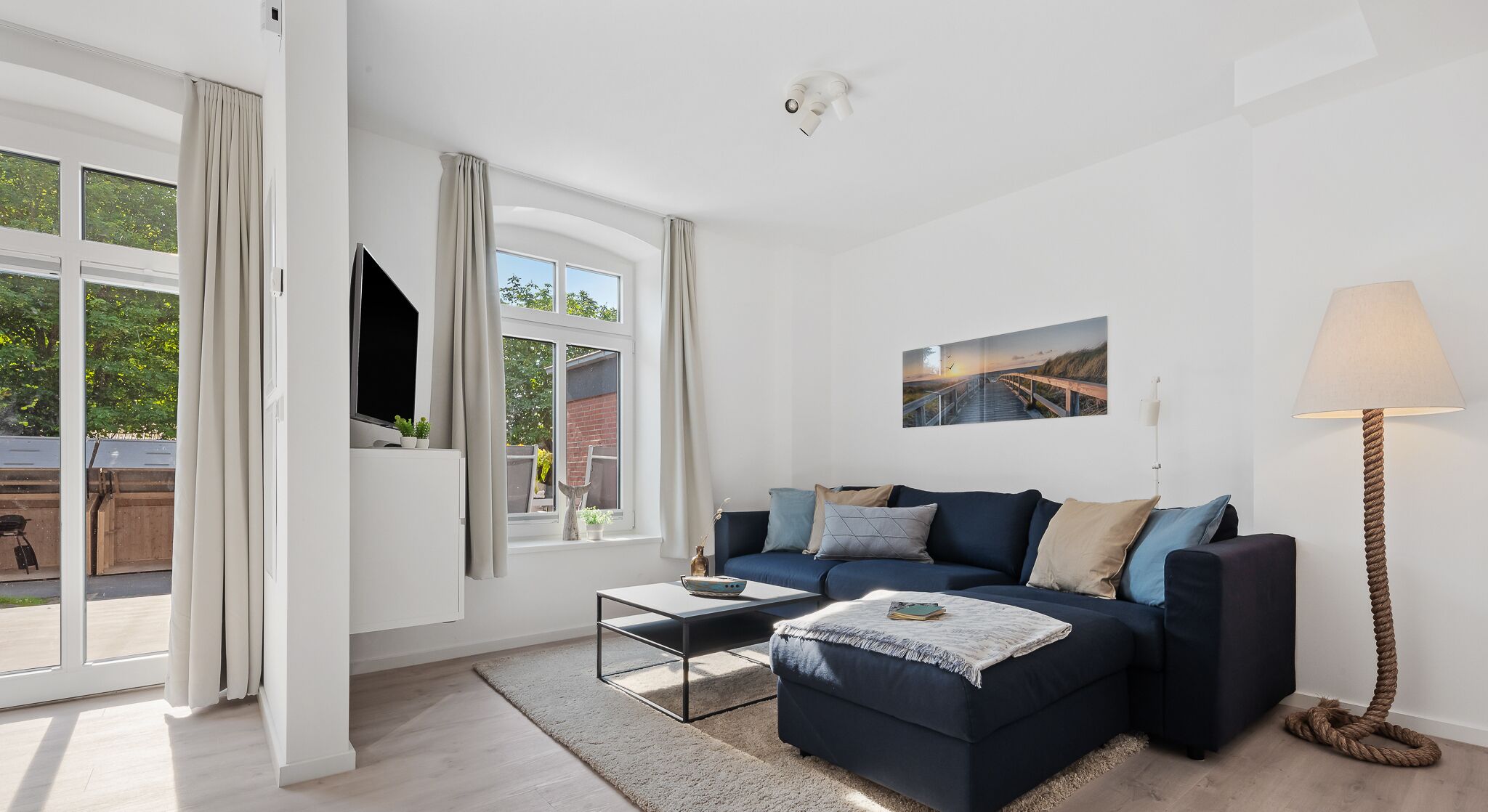 Een moderne woonkamer met een donkerblauwe bank, een salontafel en een lamp. Er is een groot raam met gordijnen, een flatscreen-tv en een ingelijste foto aan de muur.