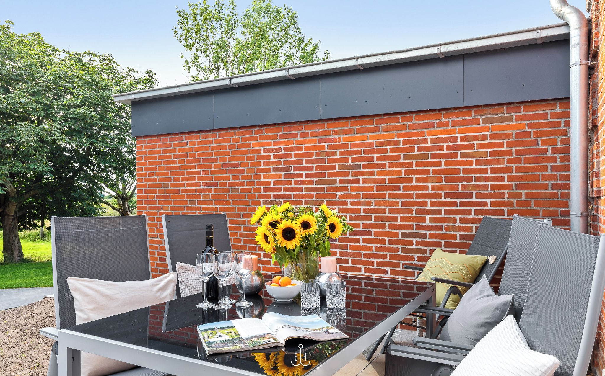 An outdoor patio with a glass table, two chairs, a vase with sunflowers, and a brick wall in the background.