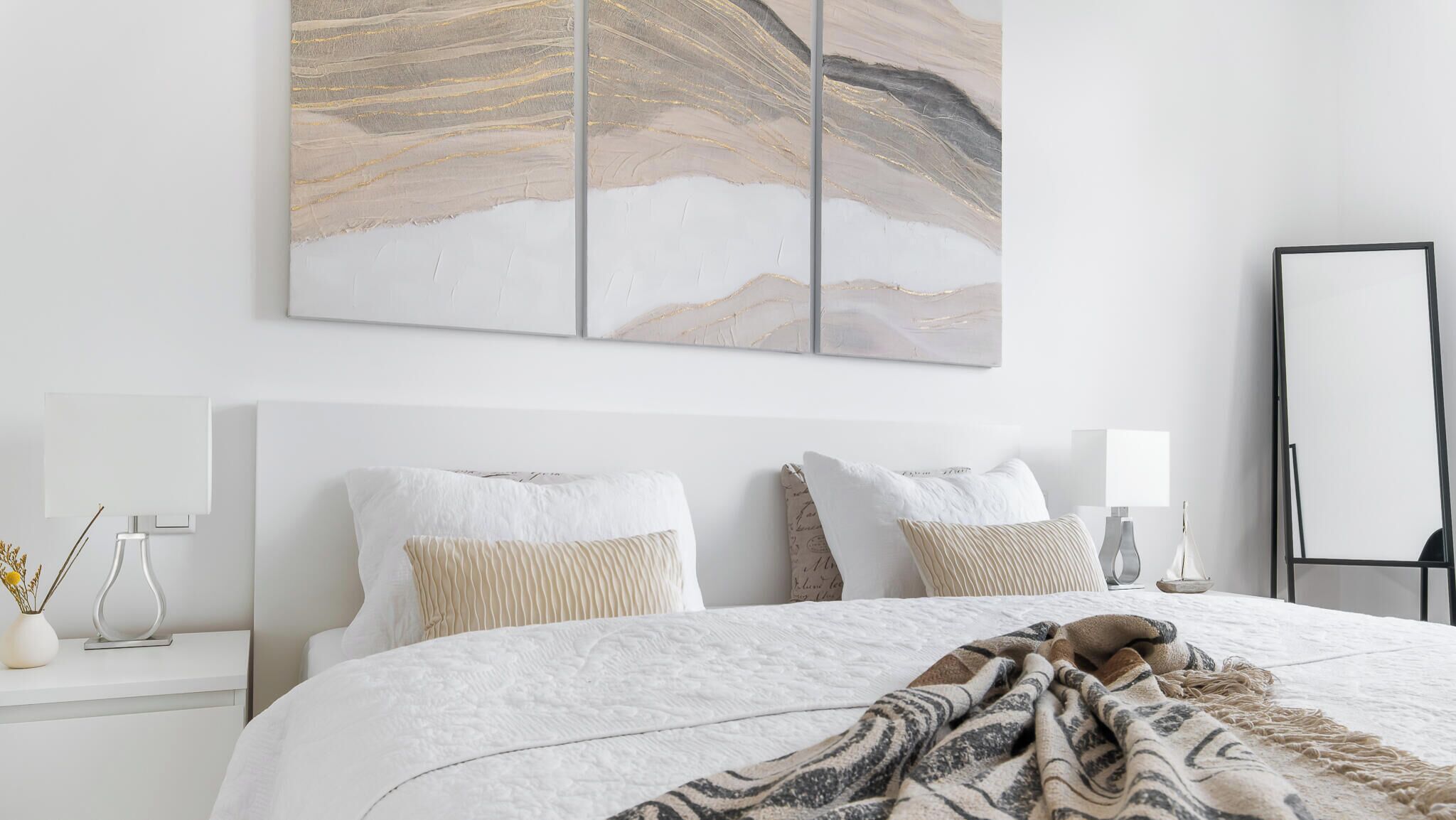 Een netjes opgemaakte slaapkamer met een groot bed, een nachtkastje met een vaas en kaars, en een spiegel aan de muur.