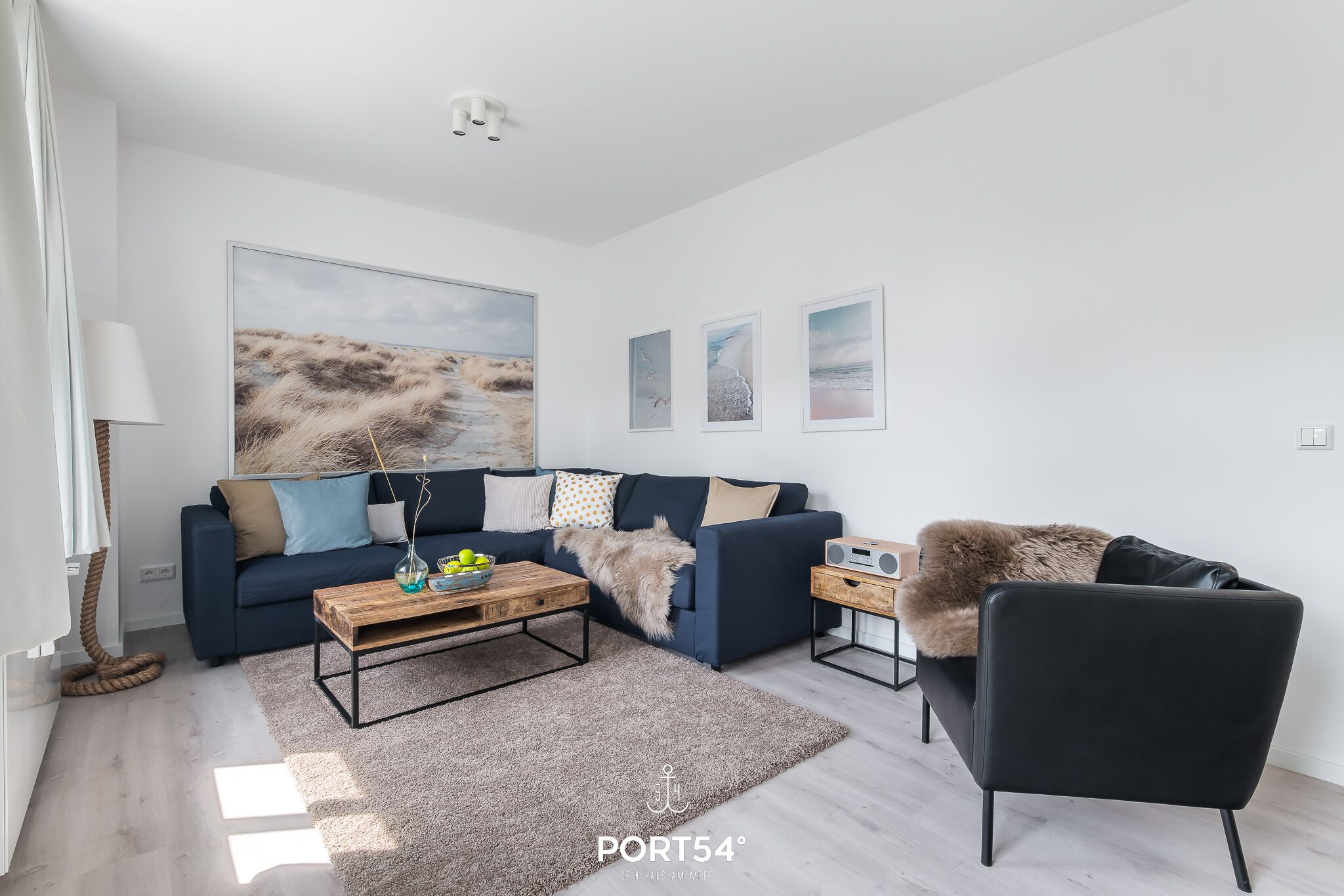 A modern living room with a dark blue sofa, a wooden coffee table, and a black armchair. There are three framed pictures on the wall and a large landscape painting above the sofa.