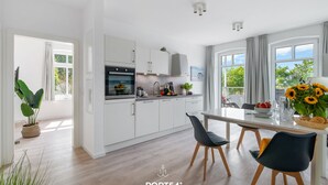 Eine moderne Küche mit weißen Schränken, einem Essbereich mit Tisch und Stühlen sowie großen Fenstern mit Vorhängen.