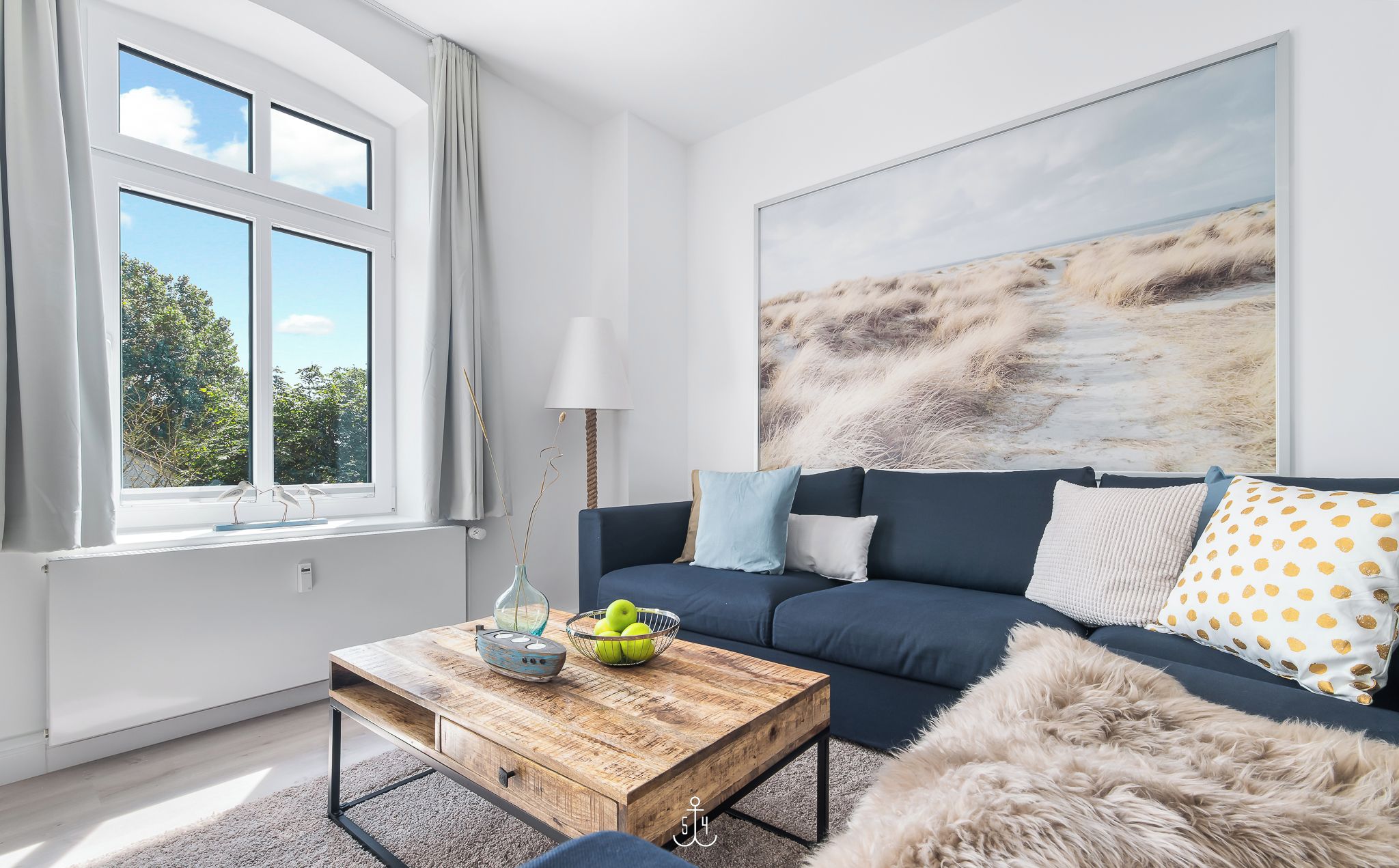 A modern living room with a large window, a blue sofa, a wooden coffee table, and a large landscape painting.