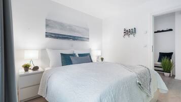 Une chambre moderne avec un lit, des tables de chevet, une commode, une étagère murale décorative et une porte donnant sur une autre pièce.