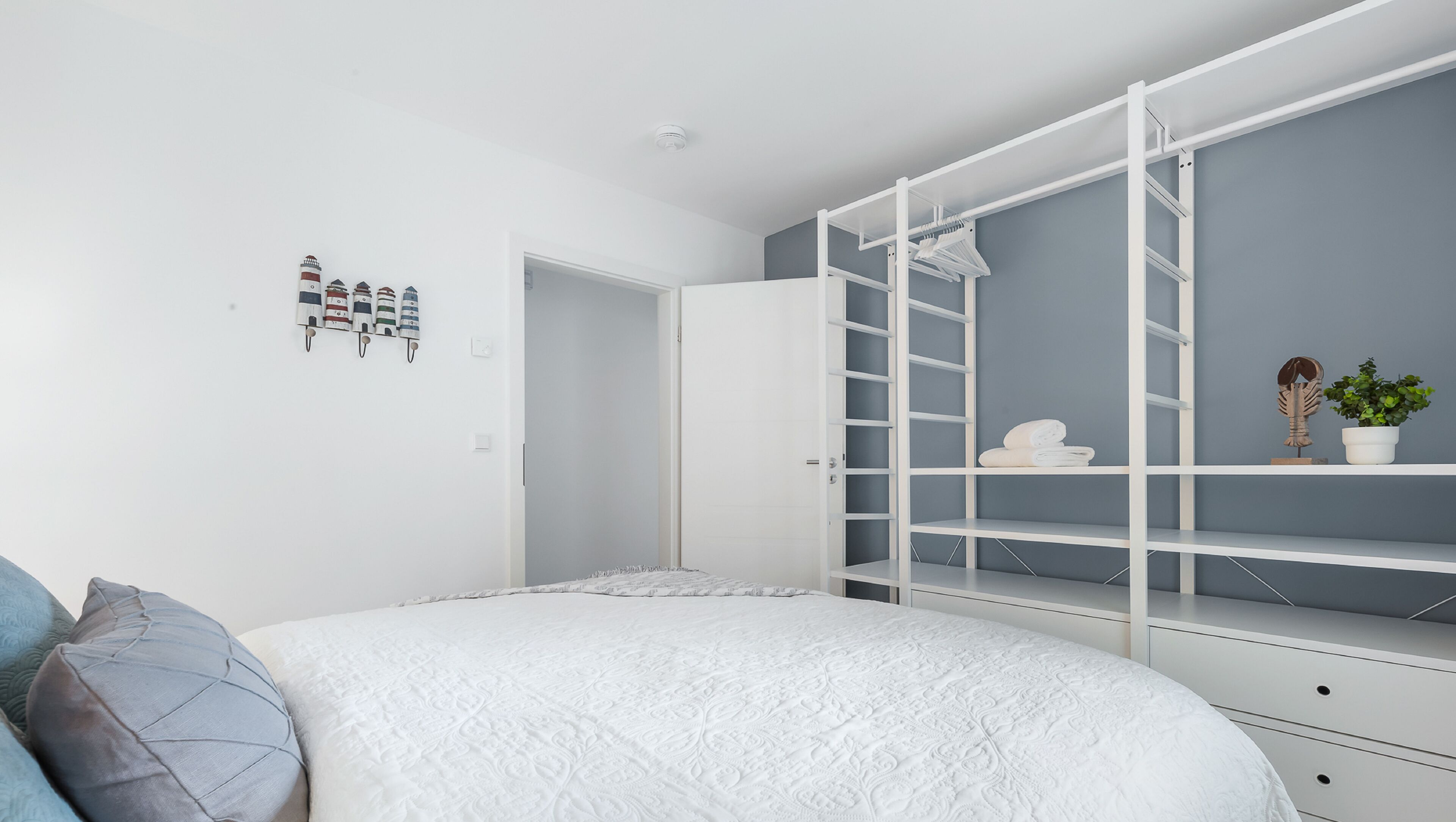 A bedroom with a bed, a white wardrobe with shelves, a small potted plant, and a wall-mounted shelf with towels.