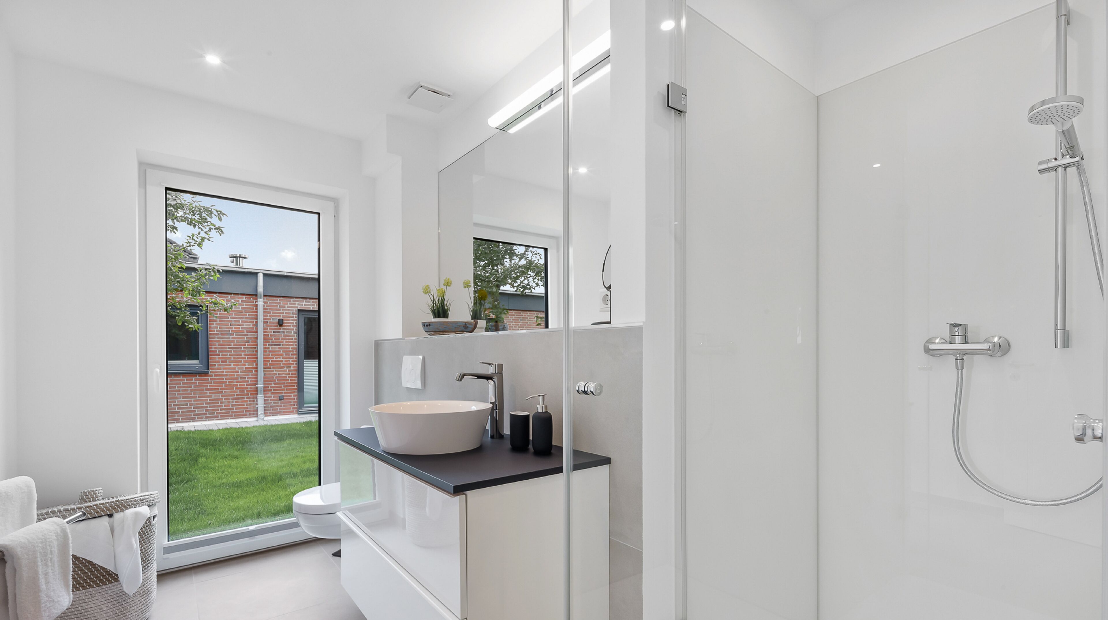 A modern bathroom with a large glass shower enclosure, a freestanding bathtub, and a large window offering a view of a brick building and greenery outside.