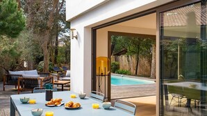 An outdoor dining area with a table set for breakfast, a pool in the background, and a glass door leading to a patio. - VILLA CHISTERA with SPA and heated swimming pool (HOSSEGOR)
