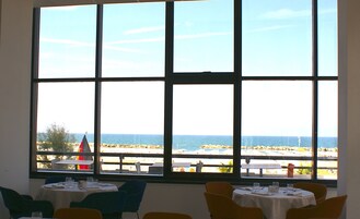 Zona pranzo con tavoli apparecchiati per due, ampie finestre con vista sul mare e sulla costa in lontananza.