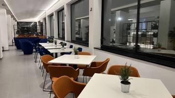 Un comedor moderno con mesas blancas, sillas anaranjadas, ventanas amplias y plantas en macetas.