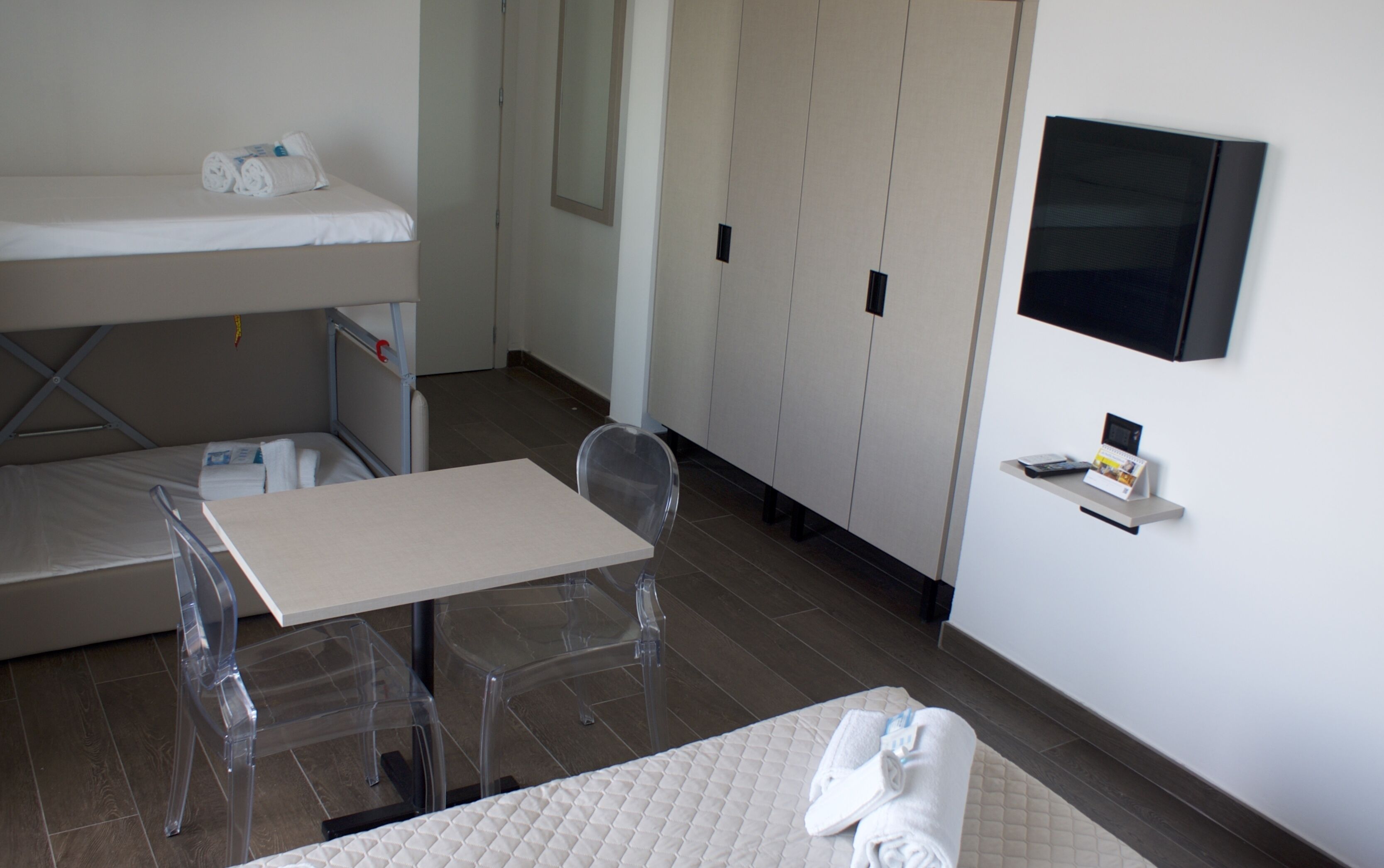 A hotel room with a bunk bed, a small table with a chair, a TV mounted on the wall, and a shelf with a book and a remote control.