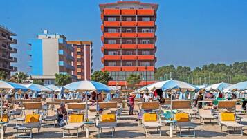 Una playa con filas de tumbonas y sombrillas, un edificio rojo alto al fondo y un cielo azul despejado.