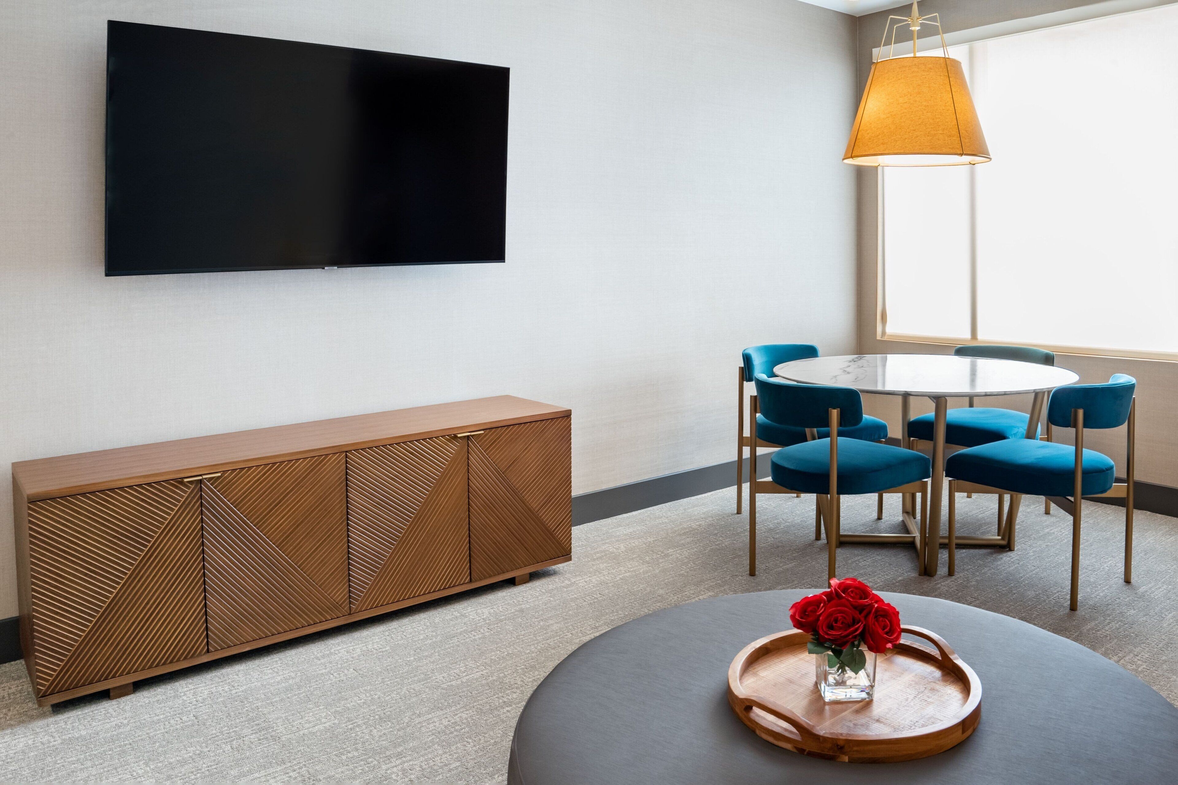 Uma sala de estar moderna com televisão de tela plana, um armário de madeira, uma mesa de centro redonda com um porta-bandejas de rosas, e uma poltrona azul disposta ao redor de uma pequena mesa.