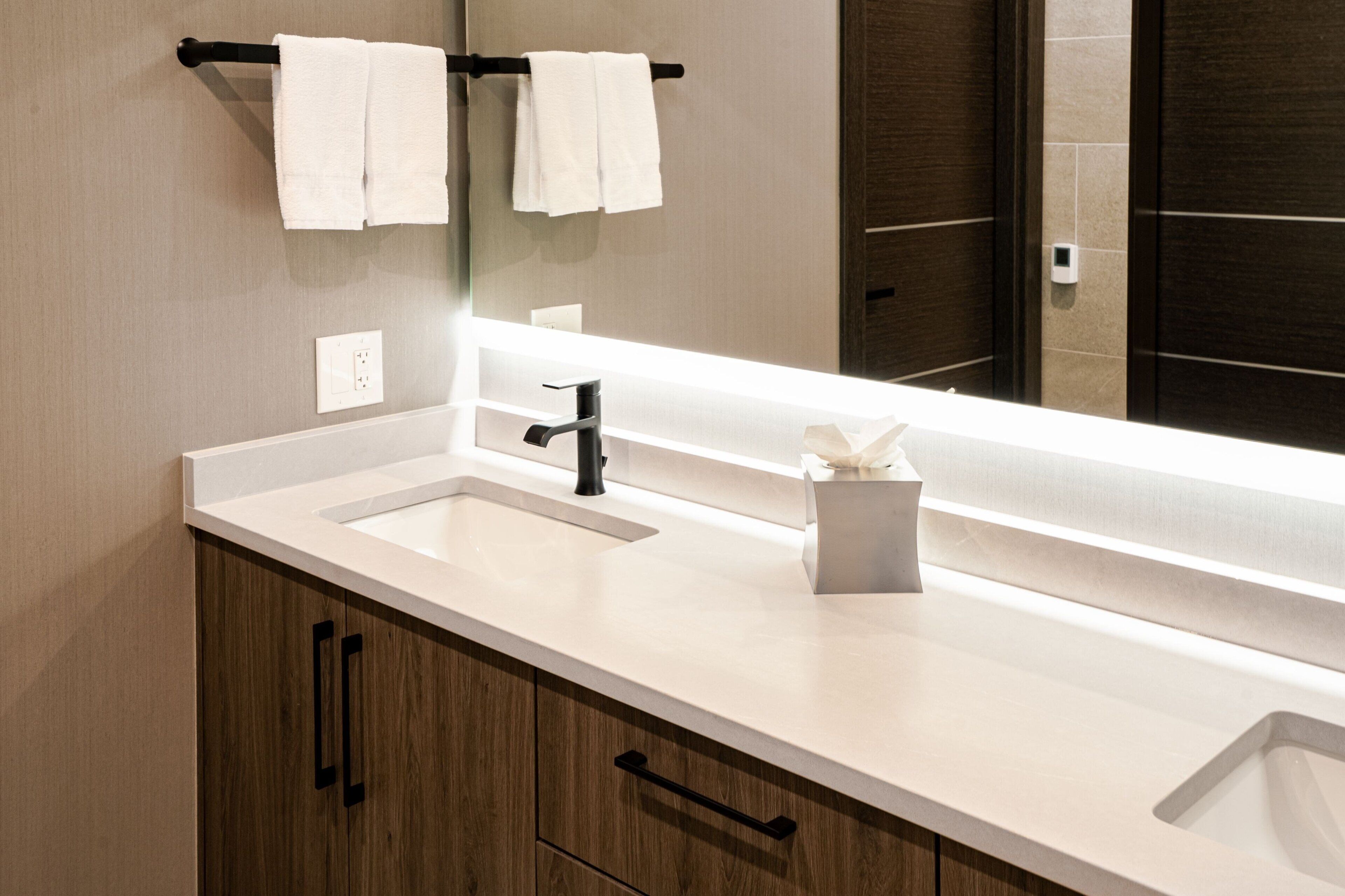 A modern bathroom with a white sink, black faucet, and wooden cabinets. There are two white towels hanging on a black towel rack above the sink.