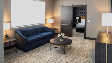 Habitación de hotel con un sofá azul, una mesa de centro redonda con un jarrón de flores, un ventanal grande y una puerta que da a un dormitorio.