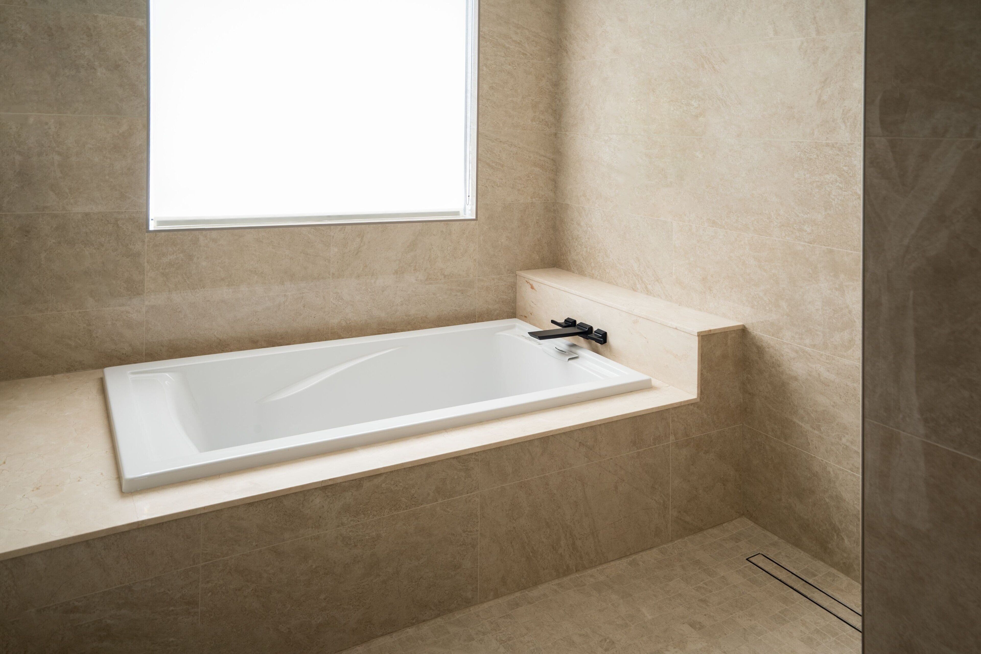 Ein modernes Badezimmer mit einer freistehenden Badewanne, einem Fenster und beige gefliesten Wänden.
