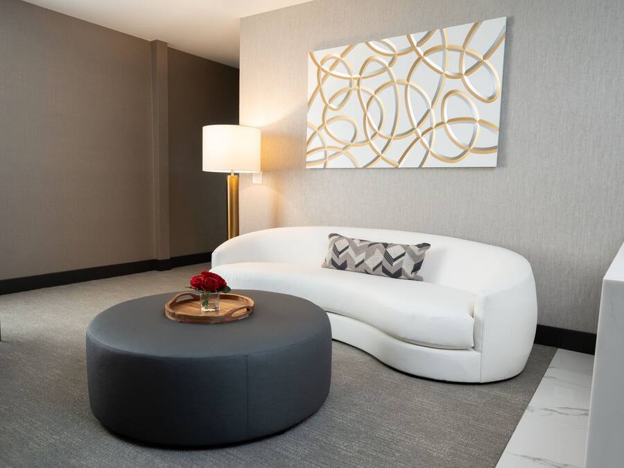 Una habitación de hotel moderna con un sofá blanco, un otoman redonda con una bandeja y una rosa, una silla azul y una obra de arte en la pared.