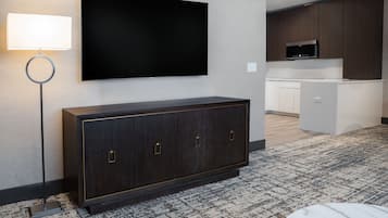 Een moderne hotelkamer met een kast van donker hout, een flatscreen televisie aan de muur, een staande lamp, een kitchenette met een magnetron, en een kamerbreed tapijt met dessin.