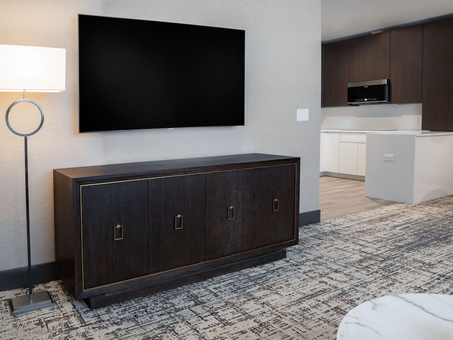 Een moderne hotelkamer met een kast van donker hout, een flatscreen televisie aan de muur, een staande lamp, een kitchenette met een magnetron, en een kamerbreed tapijt met dessin.