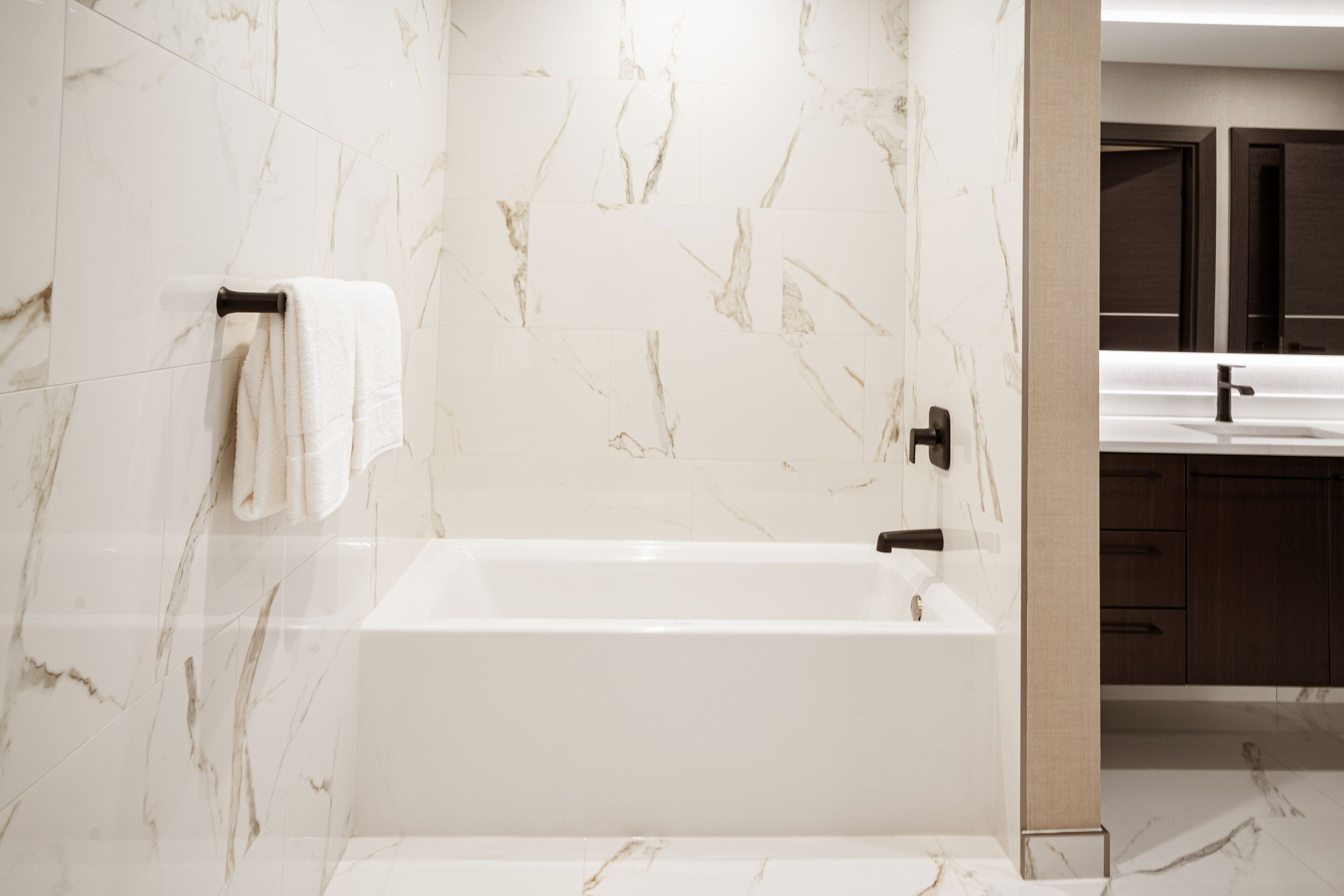 A modern bathroom with a marble wall, a white bathtub, and wooden cabinets.