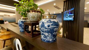 Uma área de recepção com vasos de porcelana azul e branca tradicionais sobre um balcão de madeira, uma planta em vaso e uma tela de televisão ao fundo.