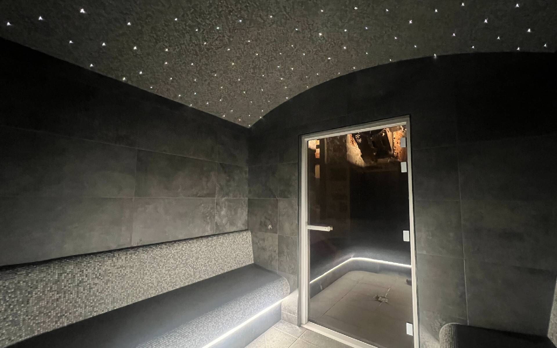 Een moderne spa ruimte met een bankje, een glazen deur naar een stoomruimte en een plafond met kleine lampjes.