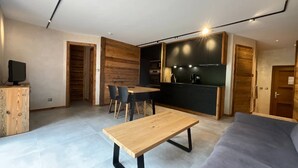A modern living room with a wooden dining table, a grey sofa, and a kitchen area with dark cabinetry and a black countertop. - Rental of 12 gîtes and 1 chalet that can accommodate 2 to 10 people. (Rochesson)