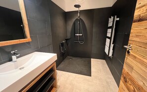 Een moderne badkamer met een witte wasbak, donkere tegels, een douche en een houten deur.