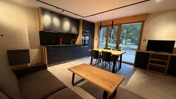 A modern kitchen with dark cabinets, a dining area with a wooden table and chairs, and a large window offering a view of the outdoors. - Rental of 12 gîtes and 1 chalet that can accommodate 2 to 10 people. (ROCHESSON)
