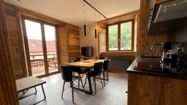 A modern kitchen with a built-in stove, a dining area with a wooden table and chairs, and large windows offering a view of the outdoors. - Rental of 12 gîtes and 1 chalet that can accommodate 2 to 10 people. (Rochesson)