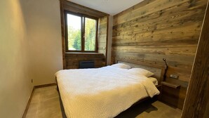 A bedroom with a large bed, wooden panel walls, a window with a view of trees, and a small bedside table. - Rental of 12 gîtes and 1 chalet that can accommodate 2 to 10 people. (ROCHESSON)