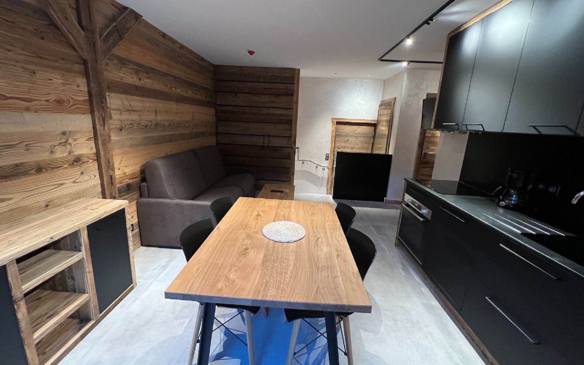 A modern kitchen with a black countertop and built-in appliances, a wooden dining table with four chairs, and a grey sofa in a corner.