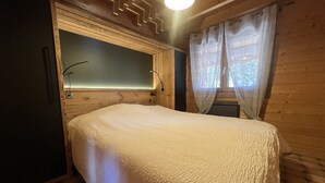 Ein Zimmer mit Holzboden, einem Bett, einem Fenster mit Vorhängen und einer Wandleuchte.