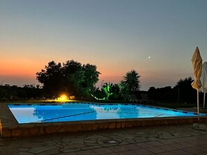 Ein Schwimmbad im Abendlicht mit Sonnenuntergang im Hintergrund, umgeben von Bäumen und einer gefliesten Terrasse.