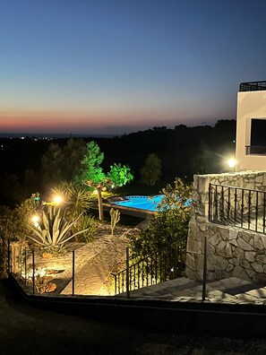 A pool area with a stone wall and metal railing, surrounded by various plants and trees. - Family-run Villa Cornelia - 3 separate apartments - tranquillity and sea views. (Rethymno - Asteri)