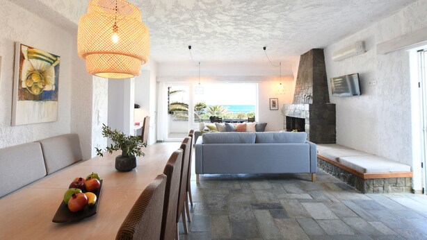 A modern living room with a sofa, dining table with fruit, a hanging light fixture, and a stone fireplace. - Villa Breath of the Sea (Hersonissos)