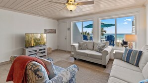 Un salon équipé d’un ventilateur de plafond, d’une télévision, d’un canapé et d’une fenêtre donnant sur la plage.