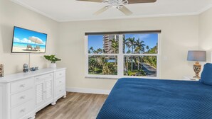 Une chambre avec un grand lit, un ventilateur de plafond, une télévision, une fenêtre donnant sur des palmiers et un immeuble, ainsi qu’une commode blanche.