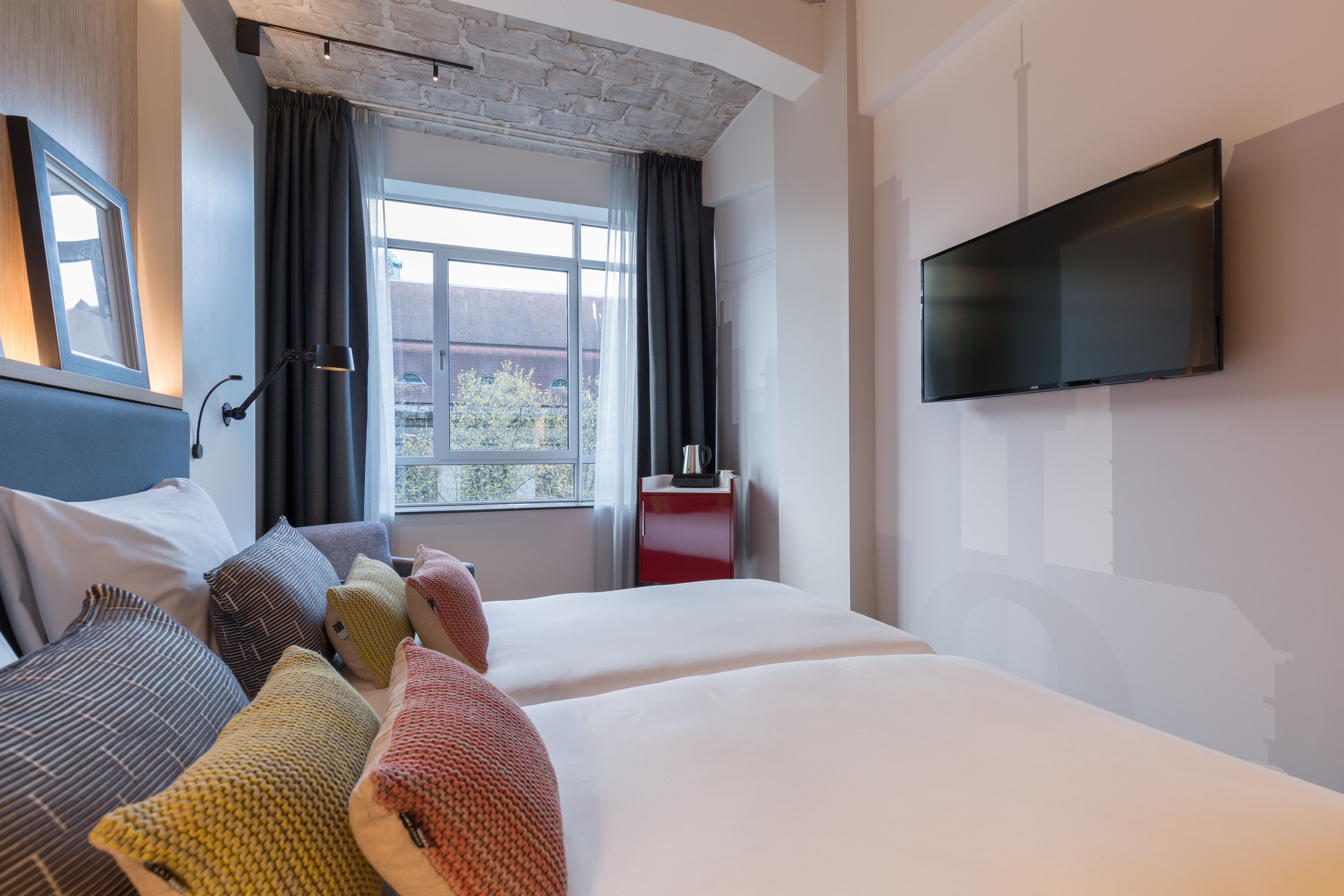 Quarto de hotel moderno com uma cama grande, televisão de tela plana, janela com cortinas e uma mesa de cabeceira vermelha.