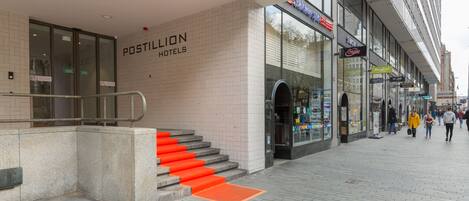 Entrada de hotel con una escalera que lleva a la puerta, un letrero que dice 'POSTILION HOTELS', y un paseo peatonal con tiendas y gente.