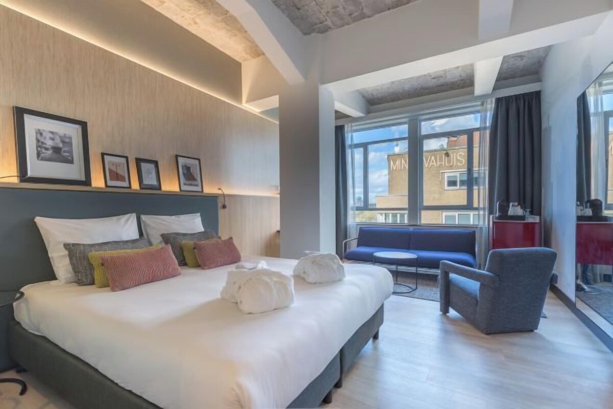 A modern hotel room with a large bed, a seating area with a blue sofa and armchairs, and a view of the city through large windows.