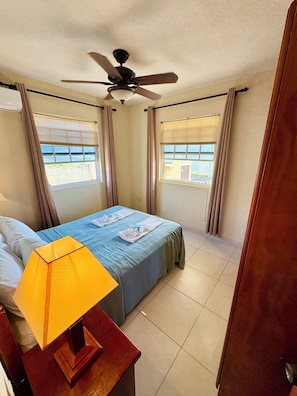 A hotel room with a bed, a ceiling fan, a window with blinds, and a lamp on a bedside table.