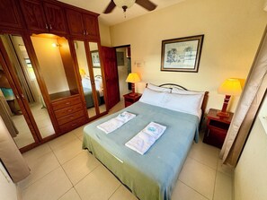 A hotel room with a bed, wooden wardrobe, two bedside lamps, a framed picture on the wall, and a ceiling fan.