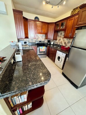 A kitchen with granite countertops, wooden cabinets, a stainless steel refrigerator, a microwave, an oven, and a washing machine.